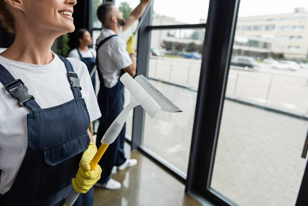 smiling woman in overalls holding window squeegee near blurred multiethnic colleagues.
